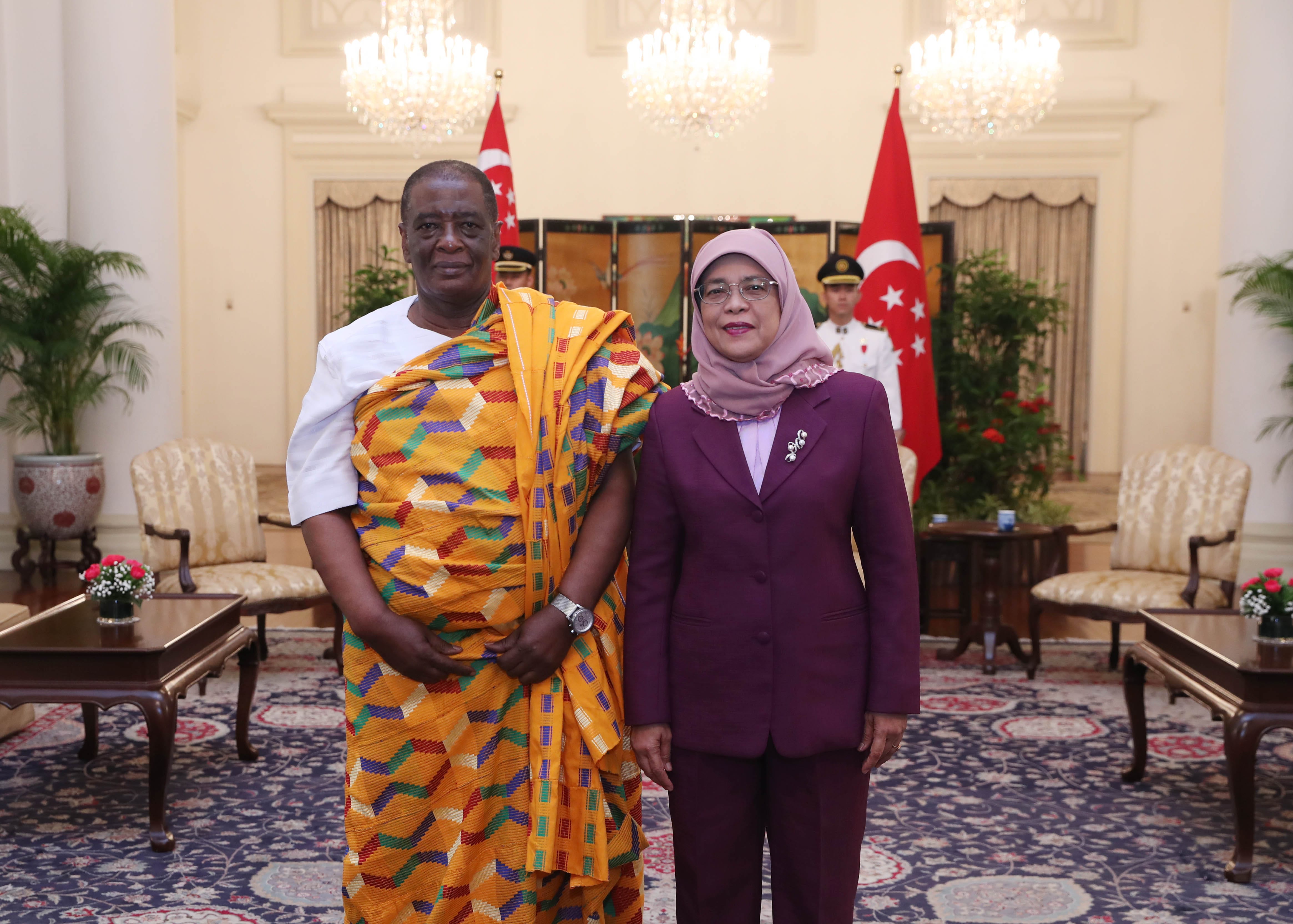 Two people standing indoors with Singaporean flags behind them. Man wears a kente cloth.
